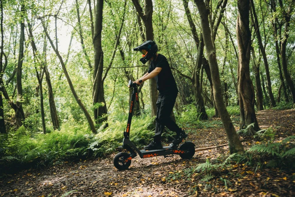 Helmeted rider navigating a woodland trail on Kukirin electric scooter, dense green forest with trees, ferns and fallen leaves, outdoor off-road adventure scene